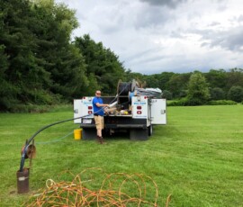 A person stands at the back of a utility truck on a grassy field, handling cables and tools. Coiled orange cords are spread on the grass, with trees and a cloudy sky in the background. A person stands at the back of a utility truck on a grassy field, handling cables and tools. Coiled orange cords are spread on the grass, with trees and a cloudy sky in the background.