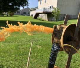 A burst of orange liquid shoots out forcefully from a valve or pipe setup on a grassy lawn, with houses and a tree visible in the sunny background. A burst of orange liquid shoots out forcefully from a valve or pipe setup on a grassy lawn, with houses and a tree visible in the sunny background.