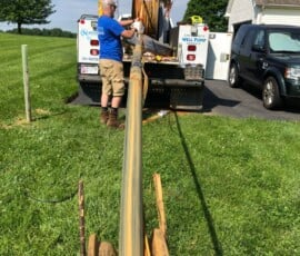 A man stands at the back of a work truck, operating machinery that is spooling a long hose or cable across a grassy yard. Tools and equipment are visible on the truck, with a house and parked SUV nearby. A man stands at the back of a work truck, operating machinery that is spooling a long hose or cable across a grassy yard. Tools and equipment are visible on the truck, with a house and parked SUV nearby.