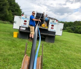 A man stands by a utility truck on grass, operating equipment that unspools black and blue cables from a reel towards the camera. The sky is cloudy and trees are in the background. A man stands by a utility truck on grass, operating equipment that unspools black and blue cables from a reel towards the camera. The sky is cloudy and trees are in the background.
