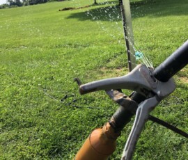 Close-up of a sprinkler head spraying water onto a green lawn, with a metal pipe and an orange attachment in the foreground. Trees and open grassy area are visible in the background on a sunny day. Close-up of a sprinkler head spraying water onto a green lawn, with a metal pipe and an orange attachment in the foreground. Trees and open grassy area are visible in the background on a sunny day.