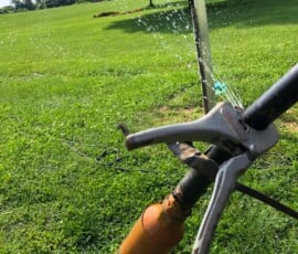 Close-up of a sprinkler head spraying water onto a green lawn, with a metal pipe and an orange attachment in the foreground. Trees and open grassy area are visible in the background on a sunny day. Close-up of a sprinkler head spraying water onto a green lawn, with a metal pipe and an orange attachment in the foreground. Trees and open grassy area are visible in the background on a sunny day.