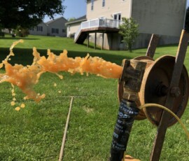 A burst of orange liquid sprays horizontally from a metal pipe apparatus set up on a grassy lawn, with houses and trees in the background under a clear blue sky. A burst of orange liquid sprays horizontally from a metal pipe apparatus set up on a grassy lawn, with houses and trees in the background under a clear blue sky.