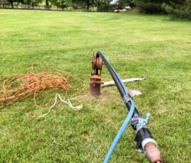 A blue cable runs through a metal pulley anchored to the ground on a grassy field, with coiled ropes lying nearby. Trees and a wooden fence are visible in the background. A blue cable runs through a metal pulley anchored to the ground on a grassy field, with coiled ropes lying nearby. Trees and a wooden fence are visible in the background.