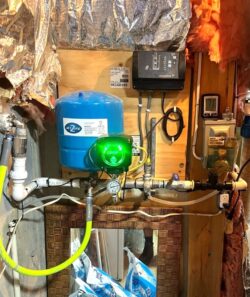 A basement wall with plumbing equipment, including a blue pressure tank, gauges, pipes, and a green-lit control box labeled DROP. Insulation and wiring are visible around the equipment. A basement wall with plumbing equipment, including a blue pressure tank, gauges, pipes, and a green-lit control box labeled DROP. Insulation and wiring are visible around the equipment.