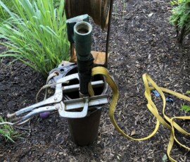 A metal tool gripping a green pipe extends from an open well casing outdoors, with a yellow nylon strap attached and soil, mulch, and plants visible around the well. A metal tool gripping a green pipe extends from an open well casing outdoors, with a yellow nylon strap attached and soil, mulch, and plants visible around the well.