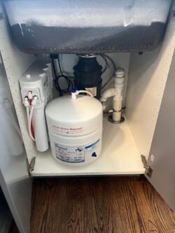 Under-sink setup with a water filtration system, storage tank, pipes, and a garbage disposal unit inside a white cabinet with open doors, set on a wooden floor. Under-sink setup with a water filtration system, storage tank, pipes, and a garbage disposal unit inside a white cabinet with open doors, set on a wooden floor.