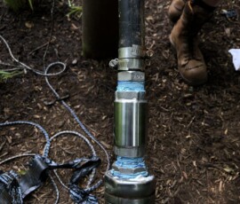 A close-up of a metal pipe joint secured with clamps and blue sealant on soil, with cables and a person wearing brown boots visible in the background. A close-up of a metal pipe joint secured with clamps and blue sealant on soil, with cables and a person wearing brown boots visible in the background.