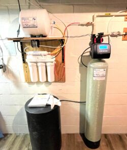 A home water filtration and softener system mounted on a basement wall, featuring multiple filter cartridges, a control unit with a digital display, a large cylindrical tank, and connecting pipes and wires. A home water filtration and softener system mounted on a basement wall, featuring multiple filter cartridges, a control unit with a digital display, a large cylindrical tank, and connecting pipes and wires.