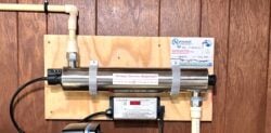 A UV water purification system mounted on a wooden wall, with pipes connected at both ends, a control unit below, and a sticker reading Annual Service Required!! on the metal chamber. A UV water purification system mounted on a wooden wall, with pipes connected at both ends, a control unit below, and a sticker reading Annual Service Required!! on the metal chamber.