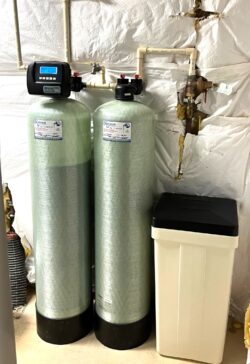 Two large cylindrical water softener tanks with digital controls stand next to a rectangular brine tank in a basement with exposed insulation and pipes on the wall behind them. Two large cylindrical water softener tanks with digital controls stand next to a rectangular brine tank in a basement with exposed insulation and pipes on the wall behind them.