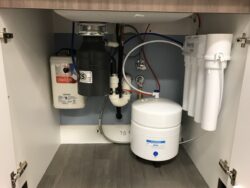 Under-sink setup with a water filtration system, a water storage tank, a garbage disposal, water heater unit, and various pipes and valves, all enclosed in a cabinet with white doors. Under-sink setup with a water filtration system, a water storage tank, a garbage disposal, water heater unit, and various pipes and valves, all enclosed in a cabinet with white doors.