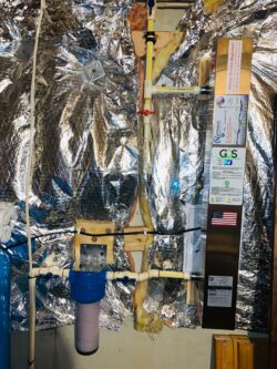 A water filtration system is attached to a basement wall with reflective insulation, featuring pipes, a blue filter housing, control valves, and a tall vertical unit with various labels and a small American flag sticker. A water filtration system is attached to a basement wall with reflective insulation, featuring pipes, a blue filter housing, control valves, and a tall vertical unit with various labels and a small American flag sticker.