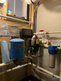 A well water filtration system mounted on a wall, featuring a blue pressure tank, water filter, various pipes, gauges, a control box, and informational signs in a utility room with exposed wooden framing. A well water filtration system mounted on a wall, featuring a blue pressure tank, water filter, various pipes, gauges, a control box, and informational signs in a utility room with exposed wooden framing.