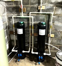 Two black water filtration tanks connected by white PVC pipes are mounted against a concrete block wall in a basement or utility room. Blue-handled valves and wiring are visible on the tanks and pipes. Two black water filtration tanks connected by white PVC pipes are mounted against a concrete block wall in a basement or utility room. Blue-handled valves and wiring are visible on the tanks and pipes.