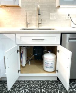 Kitchen sink with cabinet doors open, revealing an under-sink water filtration system, including filters, a tank, and connected tubing beneath a modern faucet and tiled backsplash. Kitchen sink with cabinet doors open, revealing an under-sink water filtration system, including filters, a tank, and connected tubing beneath a modern faucet and tiled backsplash.