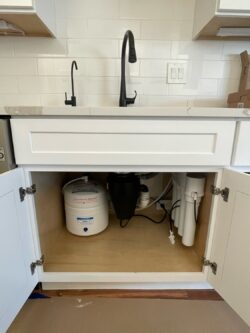 A kitchen sink with white cabinets, open cabinet doors, and a view of the plumbing underneath, including a water filtration system, water tank, and garbage disposal. White tile backsplash and black faucet are visible above. A kitchen sink with white cabinets, open cabinet doors, and a view of the plumbing underneath, including a water filtration system, water tank, and garbage disposal. White tile backsplash and black faucet are visible above.