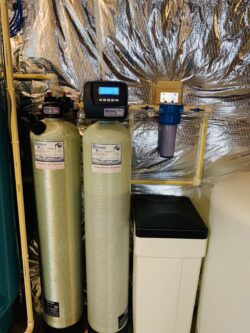 Two tall water filtration tanks, a digital control head, a blue filter canister, and a white brine tank are installed against a wall with reflective insulation in a basement or utility room. Two tall water filtration tanks, a digital control head, a blue filter canister, and a white brine tank are installed against a wall with reflective insulation in a basement or utility room.