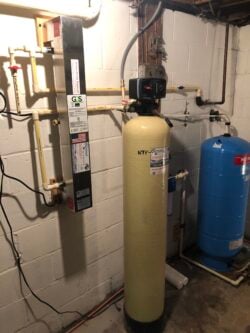 A water softener system with a tall cylindrical tank, control head, and connected pipes is installed against a basement cinder block wall. Nearby is a blue pressurized water tank. A water softener system with a tall cylindrical tank, control head, and connected pipes is installed against a basement cinder block wall. Nearby is a blue pressurized water tank.