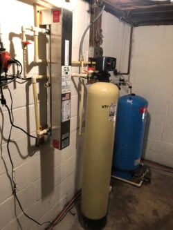 A water filtration and softener system installed in a basement, featuring a tall beige tank, a blue pressure tank, pipes, valves, and a wall-mounted control unit against a white painted cinder block wall. A water filtration and softener system installed in a basement, featuring a tall beige tank, a blue pressure tank, pipes, valves, and a wall-mounted control unit against a white painted cinder block wall.