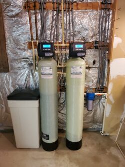 Two tall water softener tanks with digital control heads stand side by side in a basement. A brine tank is on the left, and pipes are visible on the unfinished insulated wall behind them. Two tall water softener tanks with digital control heads stand side by side in a basement. A brine tank is on the left, and pipes are visible on the unfinished insulated wall behind them.