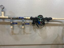 A water pipe system mounted on a wall features a pressure gauge, several blue-handled valves, and a black electronic device labeled DROP with a green light and wiring attached to PVC piping. A water pipe system mounted on a wall features a pressure gauge, several blue-handled valves, and a black electronic device labeled DROP with a green light and wiring attached to PVC piping.