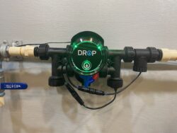 A green-lit DROP device is attached to plumbing pipes against a gray wall, with various wires and connectors visible around it. A green-lit DROP device is attached to plumbing pipes against a gray wall, with various wires and connectors visible around it.