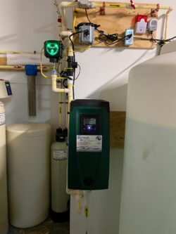 A water softener system with a green control unit, various pipes, a large cylindrical tank, and electronic controls mounted on a wall in a utility room. A water softener system with a green control unit, various pipes, a large cylindrical tank, and electronic controls mounted on a wall in a utility room.
