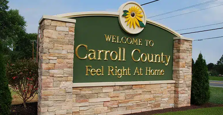 A stone sign with gold letters reads Welcome to Carroll County Feel Right At Home, topped with a yellow flower and green background—near the road and greenery where water treatment Carroll County MD ensures a fresh, clean environment.