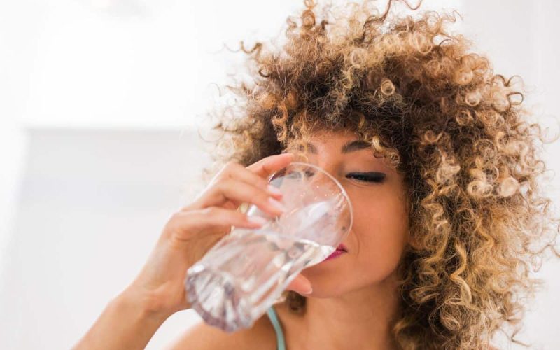 A woman with curly hair drinks a glass of water indoors, her eyes closed as she enjoys a refreshing sip—thanks to whole home water treatment systems. The background is bright and softly blurred.