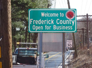 A green roadside sign reads Welcome to Frederick County Open for Business with a red emblem. Below is a speed limit 30 sign, with vehicles parked along a two-lane road—trees in the background hint at water treatment Frederick County MD initiatives.