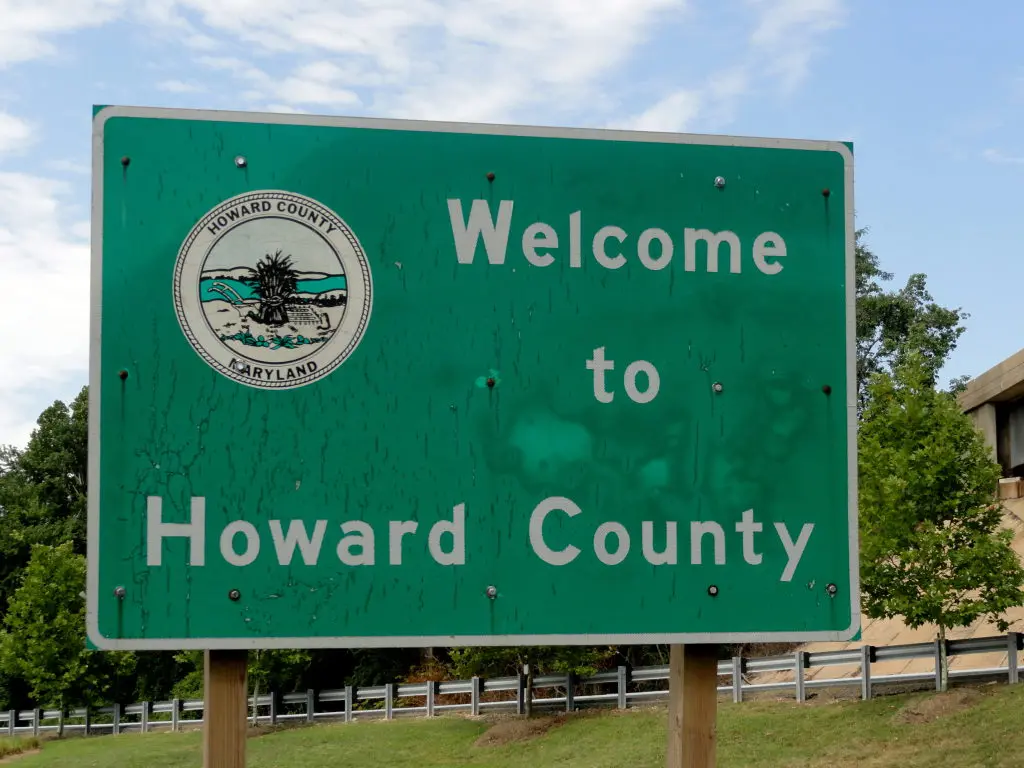 A green road sign reads “Welcome to Howard County” with the Howard County, Maryland seal on the top left. Trees and a partly cloudy sky are visible in the background—ideal for those seeking well pump services Howard County MD.