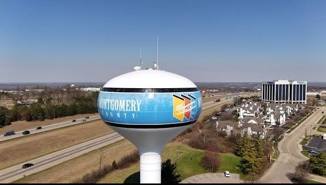 A large water tower labeled Montgomery County stands near a highway with cars, surrounded by trees and buildings under a clear sky—a reminder of the vital role well pumps Montgomery County relies on for water supply.