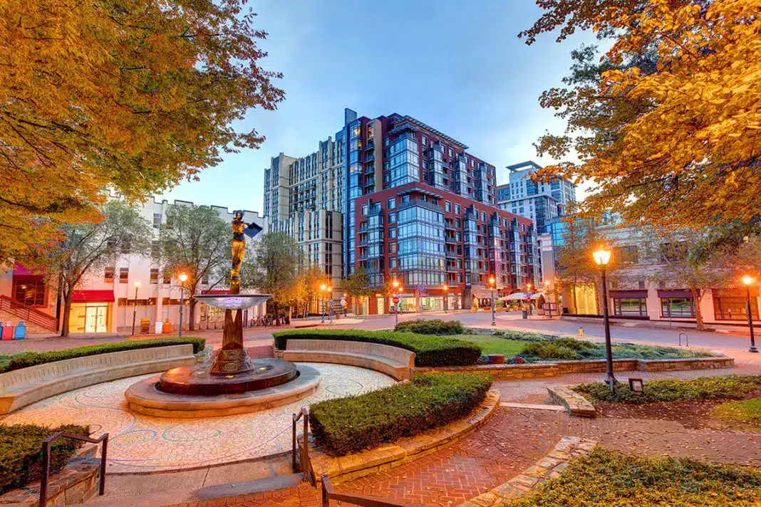 A city square with a central fountain, featuring water treatment Montgomery County MD standards, surrounded by yellow-leaved trees, landscaped hedges, brick paths, and modern mid-rise buildings under a blue dusk sky. Streetlights softly illuminate the area.