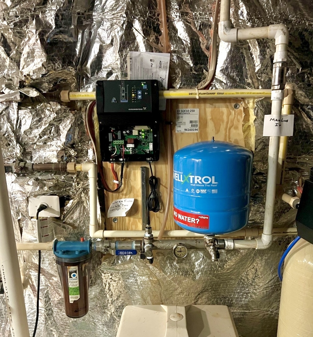 A basement utility setup with a blue Well-X-Trol water pressure tank, control panel and wiring, pipes, filter housing, and a metallic insulated wall. This Constant Pressure System for Well Water features labeled gauges on some pipes.