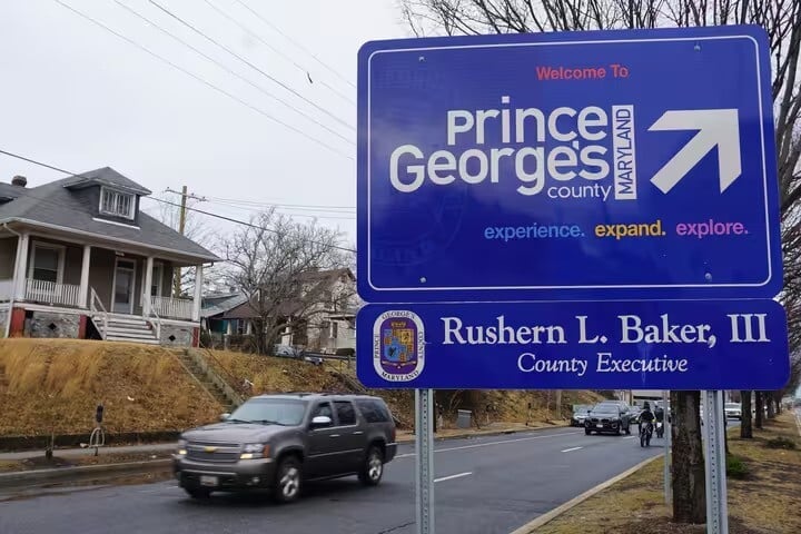 A large blue road sign welcomes drivers to Prince Georges County, Maryland, with the slogan experience. expand. explore. Cars drive by houses on a cloudy day.