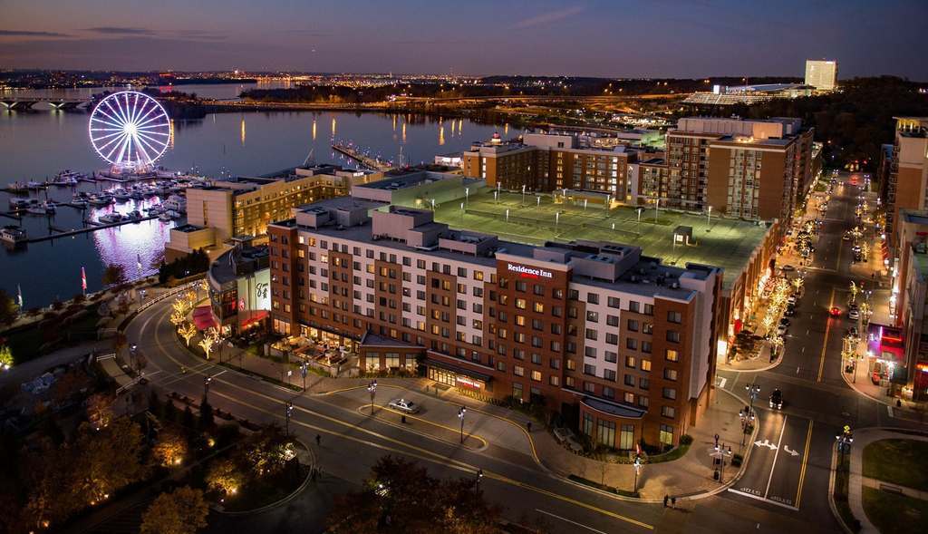 Aerial view of a city waterfront at dusk, featuring a large Ferris wheel, marina with boats, illuminated streets, and a prominent Residence Inn hotel surrounded by buildings and parking lots.