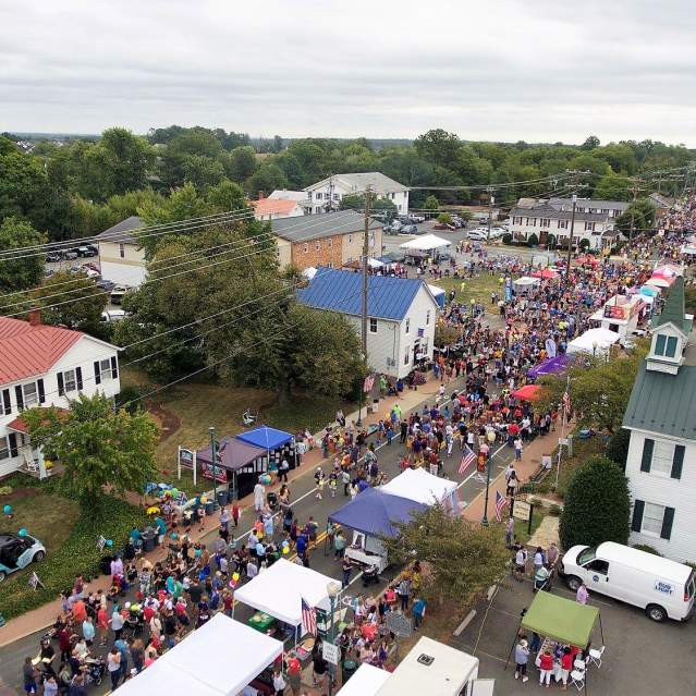 Aerial view of a busy outdoor street festival with large crowds, tents, booths, and American flags in a small town near water treatment Prince William County facilities. Houses and greenery surround the bustling event under a cloudy sky.