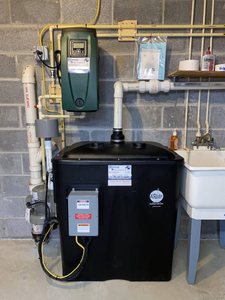 A home water filtration or treatment system for aerated water, with pipes and electrical components, is installed against a cinder block wall next to a utility sink stocked with cleaning supplies.