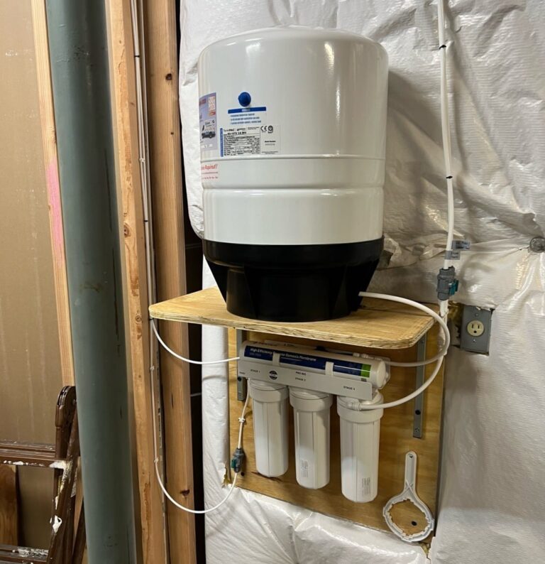 A reverse osmosis water filtration system with a large white tank and three filter cartridges is mounted on a wooden board against an unfinished basement wall with insulation and exposed framing. An outlet and white tubing are also visible.