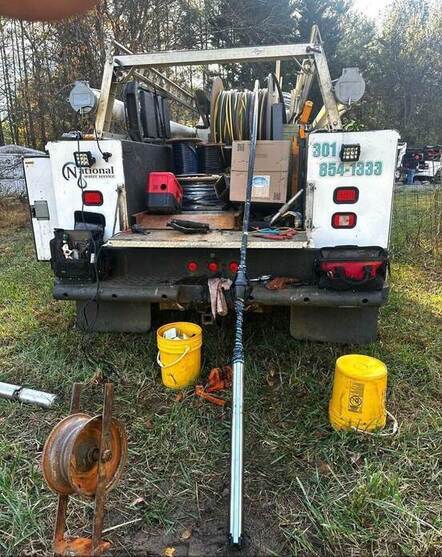 A work truck with equipment—such as hoses, cables, a submersible well pump, and boxes—is parked on grass. Two yellow buckets and a large tool sit on the ground behind it, with trees visible in the background.