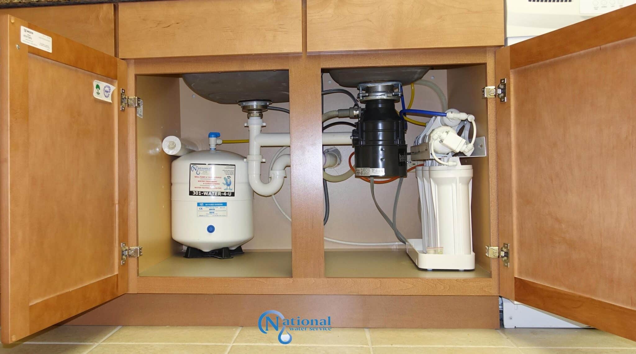 Open kitchen cabinet showing under-sink plumbing, a Reverse Osmosis water filter system, a water tank, pipes, and a garbage disposal unit. The cabinet doors are open, revealing all equipment installed inside. Open kitchen cabinet showing under-sink plumbing, a Reverse Osmosis water filter system, a water tank, pipes, and a garbage disposal unit. The cabinet doors are open, revealing all equipment installed inside.