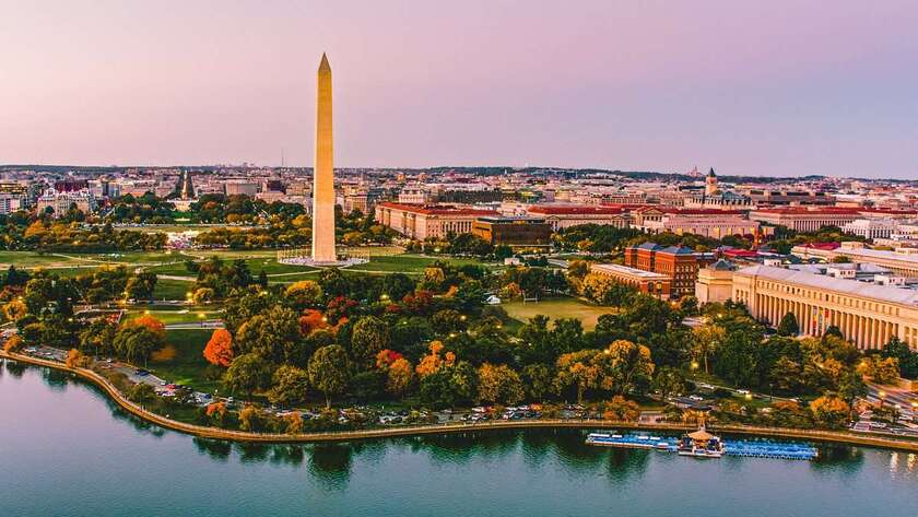 Aerial view of the Washington Monument in Washington DC, surrounded by trees, museums, and water, with city buildings in the background under a pinkish sky.
