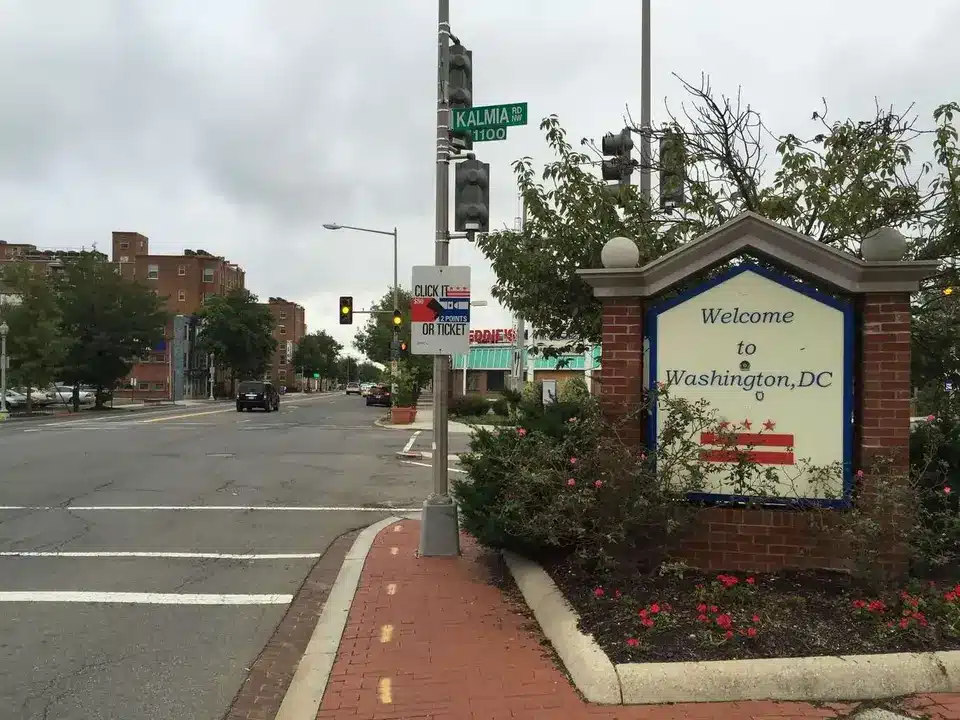 A street corner in Washington DC features a Welcome to Washington, DC sign surrounded by flowers and bushes. Nearby, a street sign reads Kalmia Rd NW. The quiet street has a few buildings and visible traffic lights.